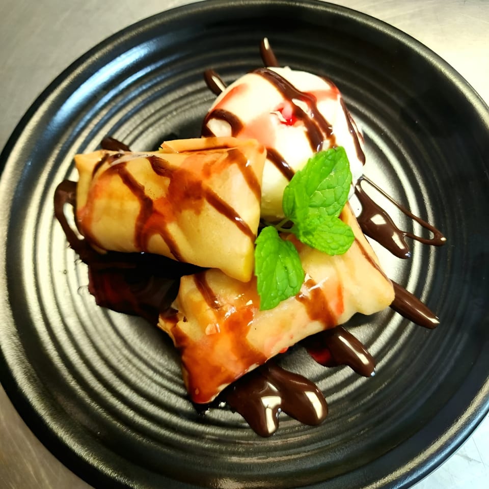 Fried banana spring rolls with ice cream, chocolate, strawberry syrup, and mint on a black plate.